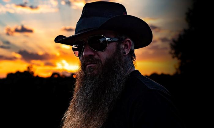 Cody Jinks wears a dark hat with matching sunglasses with the sunset as a backdrop. 