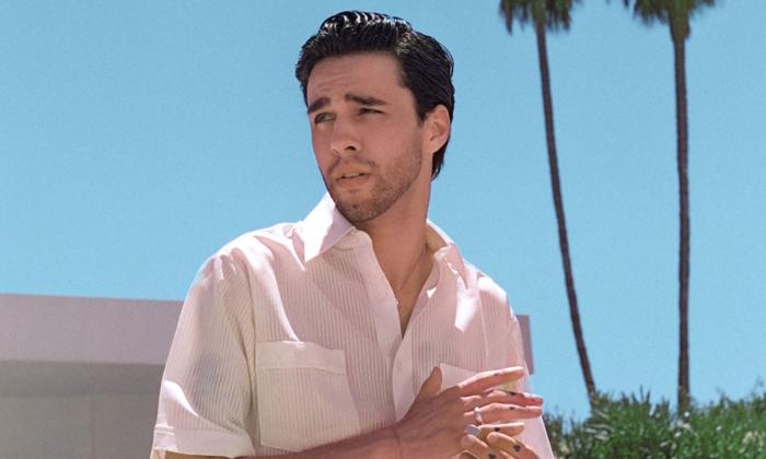 Stephen Sanchez wears a white dress shirt and poses in front of two palm trees. 