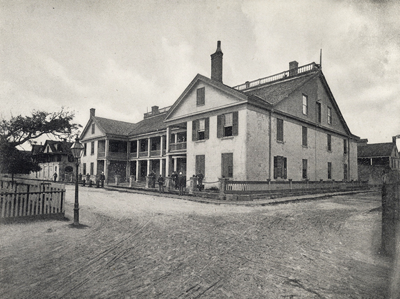 A black and white photograph of a building in St. Augustine, Florida. TIn the foreground is an intersection of sandy streets. The building that is the focus of the image is a three story building that nearly extends down the entire block. Several soldiers in uniform stand in the front of the building. 