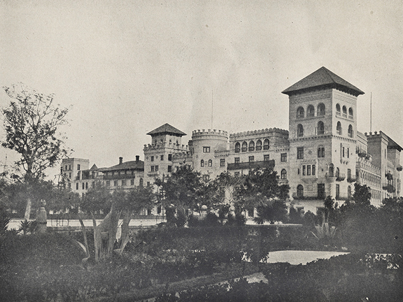 A historical photograph in black and white of the Cordova Hotel (now the Casa Monica)in St. Augustine, Florida. It is a grand hotel with many different architectural features in the Spanish style. In the foreground of the photo, the street and sidewalk in front of the Cordova is hidden by the gardens of the Hotel Alcazar (now the Lightner Museum) across the street.