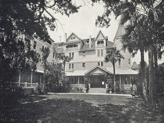 A black and white photograph of an enormous hotel in St. Augustine, Florida. It is a four story building with several chimneys, dozens of windows, and a wrap around porch. On the right of the photo, the building has a tall spire. A lawn stretches into the foreground and the image is framed by oak trees and palm trees, which throw shade on the grass.