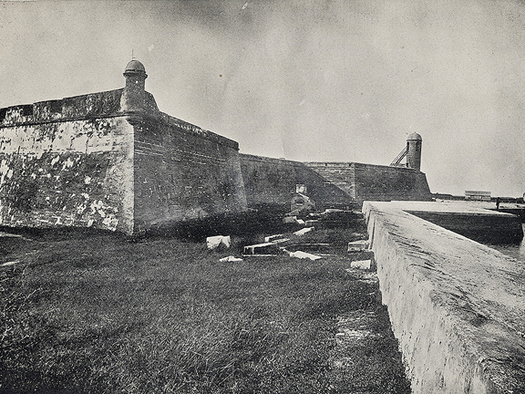 A black and white photograph of the Castillo de San Marcos (then Fort Marion) in St. Augustine Florida. The photo faces north and features the sea wall along Matanzas Bay on the right side of the image. The coquina for stands in a 'moat' full of grass.