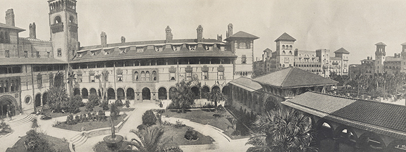 A historical photograph in black and white. Panorama view of the courtyard of the Hotel Ponce de Leon in St. Augustine, Florida. The courtyard is criss-crossed with coquina stone paths and filled with tropical plantlife.