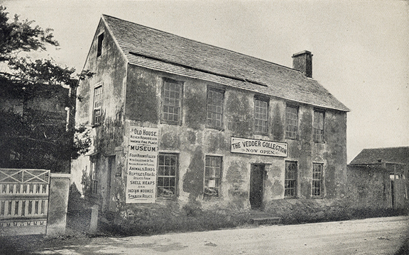 A historical photograph in black and white of the Vedder Museum in St. Augustin, Florida (which no longer exists). It is a two story house with an attique, seemingly made of coquina, as it is crumbling on the outside. The street in the foreground is sand.