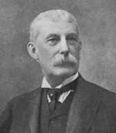 A black and white photograph of Henry M. Flagler from the chest up. He is old in this image, with short white hair parted in the middle and a bushy white mustache. He wears a three-piece suit and a pearl in his tight tie.