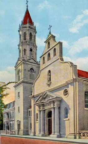 A color-tinted photograph of the Cathedral Basilica of St. Augustine (then only the Cathedral of St. Augustine), circa 1920s. It is a Spanish-Mission style building with a red roof and white plaster exterior. Facing West, the facade of the cathedral holds four bells and the image of St. Augustine. The bell tower stands on the left side of the photograph.