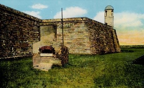A color-tinted photograph of the Hot Shot Oven at the Castillo de San Marcos (then Fort Marion) in St. Augustine, Florida. The oven is a cement construction with a semi-circular opening meant to deposit cannon balls. The stone bastions of the fort are in the background. The sky is blue with a yellow tint on the horizon. The fort lawn is tinted bright green.