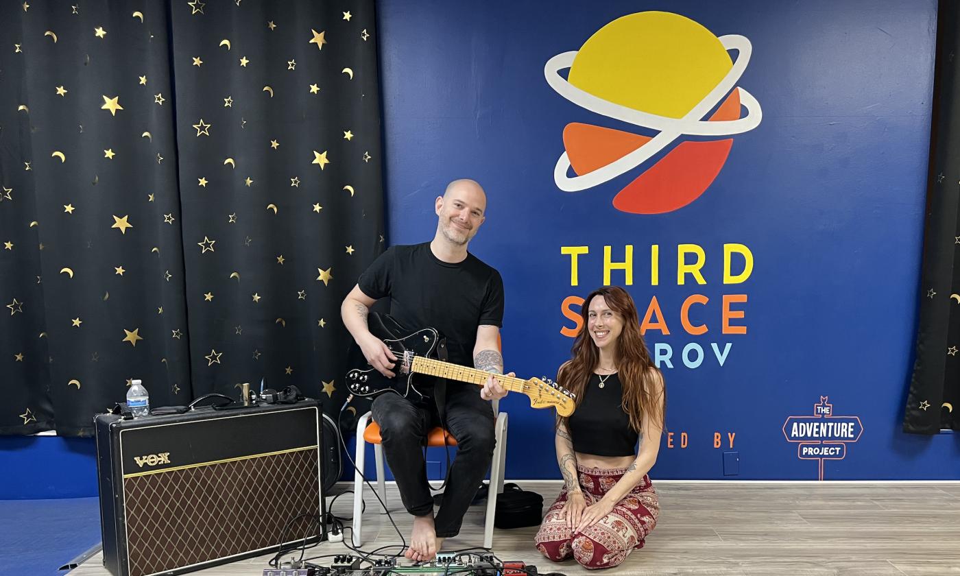 A man with a guitar and amp sitting on a chair next to a woman on the floor in her yoga clothes