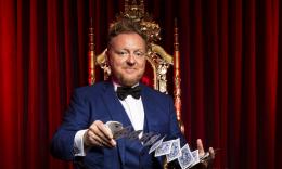 Magician Bill Abbott in a blue tux, performing a card trick in front of red velvet curtains