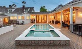 The pool deck off this L-shaped home shows a pool and hot tub with a warmly lit deck in the evening