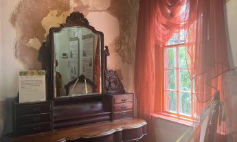 An antique dark wood vanity next to a window with gauzy red curtains and sun streaming through.