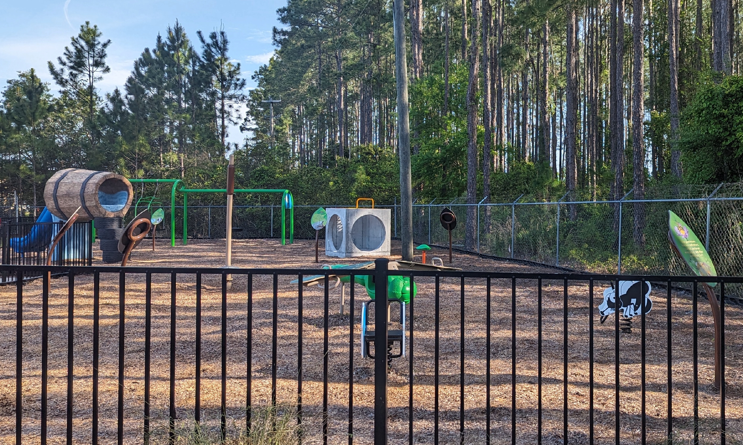A playground near a stand of trees, is fenced in and covered in wood mulch