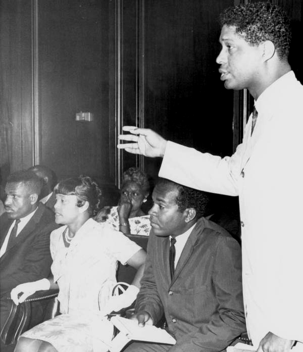 A black and white photo. Dr. R.B. Hayling stands in the foreground, in the middle of speaking intensely. Behind him, a group of fellow Civil Rights Activists sit, looking out of frame.
