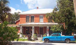 A photograph of a two story house in St. Augustine, Florida. The house is bright orange in the sunlight, and surrounded by palm trees and oak trees. There is a porch facing the street with white pillars. A blue pick up truck is parked out front.