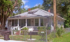 A photograph of a home in St. Augustine, Florida. It is a beige house with white accents and a dark grey shingled roof. The roof has a dormer window, suggesting a second floor. There are also two red chimneys visible above the roof. The yard, which is fenced in by chain link, is bright green with bushes and grass.