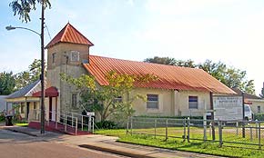 An image of a large church building in St. Augustine, Florida's Lincolnville neighborhood. The church has a red tin roof with a yellow brick structure. Facing the street, a rectangular-prism shaped tower stands. At the base of the tower, a red awning marks the front door of the Bethel Baptist Church.