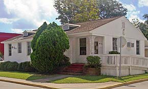 A photograph of the Rudcarlie building in St. Augustine, Florida. It is a whit building with a brown shingled roof. The steps are painted red and there is a small cement path connecting the house to the sidewalk. The picture was taken on a sunny day and the plants and grass are a bright green. There is a wheelchair ramp on the side of the building that faces the street.