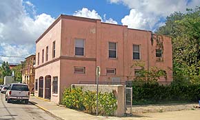 A photograph of a two story building in the Lincolnville neighborhood of St. Augustine, Florida. Taken at a three-fourths angle, the building has a pink stucco facade and rows of windows. A dirt parking lot is in the foreground and there are cars parked on the street, on the lefthand side of the image.