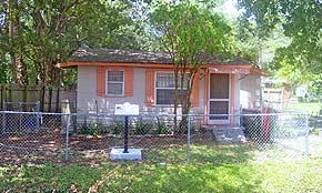 A sunny photograph of a house in St. Augustine, Florida.The house is wooden and beige with orange accents. Trees surround and shade the yard. The ACCORD Freedom Trail marker plaque faces the street near the chain link fence.