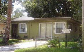 A photograph of a one-story house in St. Augustine, Florida. The house is pea green with white windowsills and accents. To the right of the sun-soaked driveway, the ACCORD freedom trail plaque for the site can be seen in the grass.