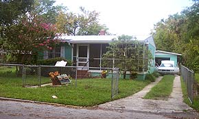 A photograph of a turquoise colored house in St. Augustine, Florida. The sunny yard is filled with plants and there is a white car in the dirt driveway. The ACCORD Trail plaque can be seen behind the chain link fence in the yard.