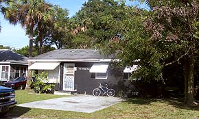 A photograph of a one-story house in St. Augustine, Florida. It is a dark grey house with white awnings and accents. It is surrounded in palm and oak trees.