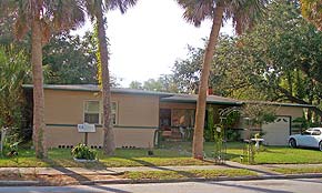 A photograph of a one story Ranch style house in St. Augustine, Florida's Lincolnville neighborhood. Palm trees line the sidewalk facing the street, and between them the ACCORD Freedom Trail marker stands. The house is tan with a flat shingle roof and there is a white car in the driveway, which is on the right hand side of the image.