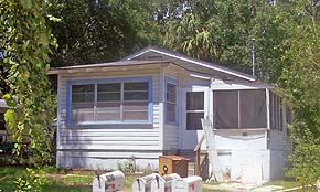 A photograph of a white, one story house in St. Augustine, Florida. The house has a screened in porch on the right side of the image. Its window frames are painted light blue. The background is filled with Florida foliage of palm trees and oak trees.