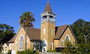 A photograph of a church in the Lincolnville neighborhood of St. Augustine, Florida. The sky is a perfect blue and the greens of the palm and oak trees are bright. The church is made of a yellow-toned brick, with white window frames and a dark brown shingled roof. A tower shaped like a rectangular prism is in the middle of two wings of the church, and has a pyramid shaped roof above an open section that holds the church bells.