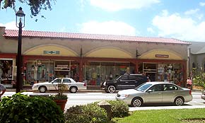 A photograph of several storefronts on King Street in St. Augustine, Florida. Taken from across the street, there are cars parked on both sides. The building has a brick red roof and large store windows are framed with brickwork on the exterior.