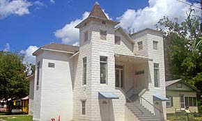 A sunny photograph of a white church in St. Augustine, Florida. The church steps face the camera, and lead up to a covered porch. On either side of the porch are two towers — one with a pyramid-shaped roof, and other with a flat roof.