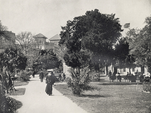 A historical image in black and white. It shows a woman in a long dark dress walking along a paved path next to tropical shrubbery.