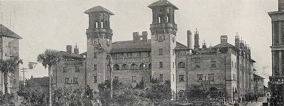 A historical photograph in black and white. A view of the Hotel Alcazar (now St. Augustine City Hall and the Lightner Museum) in St. Augustine, Florida. The Alcazar has two main towers on its King Street facing wall, and a courtyard of palmetto trees in the front. The view down Granada Street reveals pedestrians and carriages.