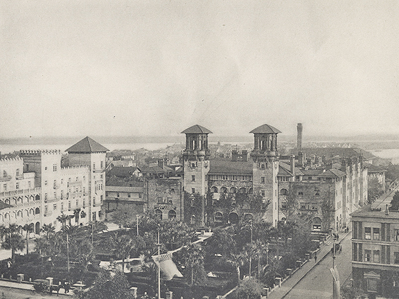 A historical photograph in black and white. An aerial view of the courtyard of the Alcazar hotel (now the Lightner Museum). The courtyard is populated with huge palm trees. Granada street on the right side of the image has one sole pedestrian.