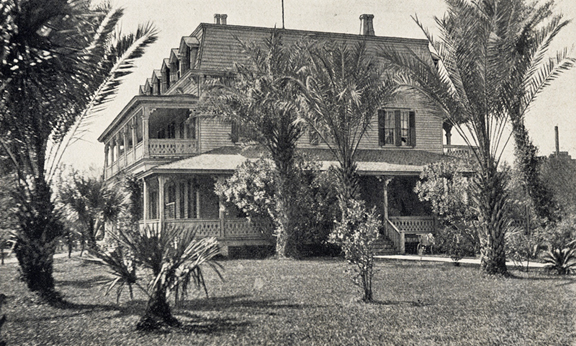 A black and white photograph of a large house in St. Augustine, Florida. It is a three story house of wood with a panel facade. Its ground and second floors have balconies white the third has several dormer windows. At least three chimneys stand above the roof. The foreground is filled with a lawn that has many tropical plants-- mostly palm trees. 