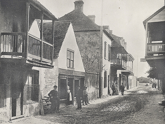 A black and white photograph of a street in St. Augustine, Florida. The focus is on the left side of the image, where a line of buildings can be seen. They are mostly two-story buildings made of coquina stone or stucco. Some have balconies that overhang the muddy street. In the foreground, some men in dark suits and hats face the camera, the closest ot the camera having a mustache. Further down the road, a few more people face the camera, women included. All of these people seem to be White. 