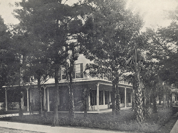 A black and white photograph of a large house in St. Augustine, Florida. It looks to be at least three stories but is shrouded by palm and cedar trees, which line the sidewalk. The house has a wrap around porch that is dotted with chairs. The figure of a small person can be seen on the walkway to the house on the left of the image. 