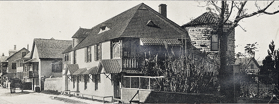 A historical photograph in black and white. View of a house on St. Francis Street in St. Augustine, Florida. It is a two story house with an odd stone tower in the bacl. 