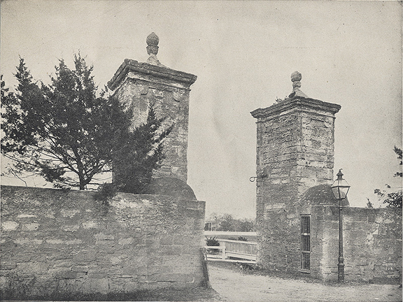 A black and white photograph of the Old City Gates of St. Augustine, Florida. This historical image was probably taken in the early 1900s. The gates are made of blacks of coquina stone and have two square towers on either side of a dirt road and a red cypress tree can be seen on the left side of the image, growing taller than the walls of the gate.