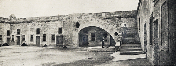 A historical photograph in black and white. An interior view of the Castillo de San Marcos (then Fort Marion) that shows the Eastern staircase to the lookout deck and Eastern wall of the fort, which has a line of doors and windows to the inner chambers.