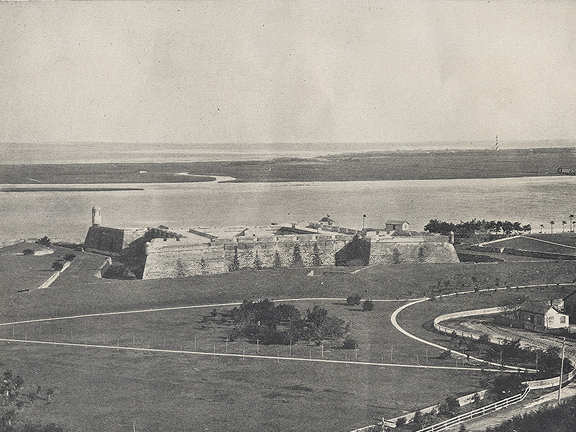 A historical photograph in black and white. Aerial view of the Matanzas Bay and the Castillo de San Marcos (then called Fort Marion). The foreground is filled with the fort lawn, which is criss-crossed with white sand walking paths. The fort is a coquina structure with a bastion to the North (left side of the image). The photo is arranged so that Anastasia Island is visible in the background, as well as the Atlantic Ocean. The St. Augustine Lighthouse is also visible on the right side of the image.
