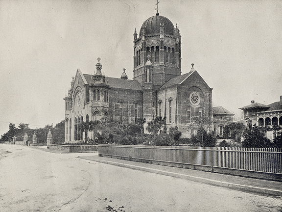 A black and white photograph of a grand church in St. Augustine, Florida. In the foreground is a sandy street and a small pile of horse droppings. A fence extends from the right hand side of the image towards the focus of the photo, which is a massive church. The church is made of stone and stands at well over three stories tall, the tallest structure of the church being a domed tower that has a cross on top. Palm trees being grown on the ground are dwarfed by the beautiful building's spires and windows.