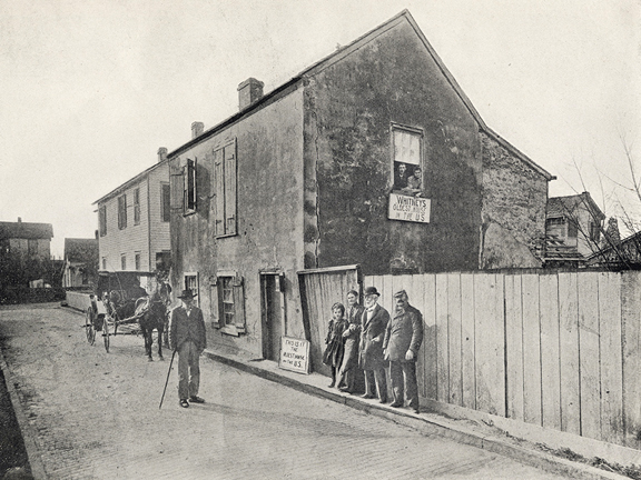 A black and white photograph of a street in St. Augustine, Florida. In the foreground on the right side of the image a group of white people stand against a fence -- two men, a woman, and a child. More white people stand in the street near a carriage.These people are in front of a two story house that is likely made of coquina. Facing the camera, two more people (a young couple) look out the window. Underneath their window is a sign that reads "WHITNEY'S: Oldest House in the US" 