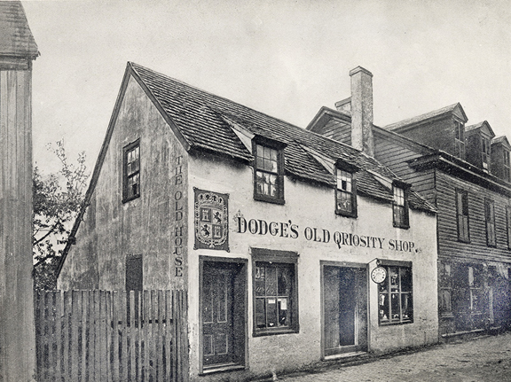 A black and white photograph of a two story building in St. Augustine, Florida. The side of the building that faces the brick street is painted with the words "Dodge's Old Qriosity Shop." in a fantastical serif font. To the left of those words is an image of Spanish coat of arms. On the side of the house that faces south, the words "The Old House" are painted in the same font. There are two doors facing the street and two windows. The windows look to have items displayed.