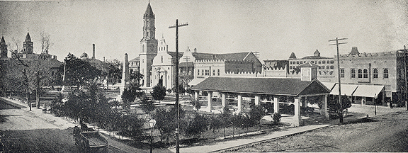 A historical image in black and white. A panoramic view of the Plaza de la Constitution in St. Augustine, Florida. The Cathedral's spire can be seen in the center of the image, with the old Public Market in the foreground on the right. Both the monument to the constitution and the old Confederate monument are visible.