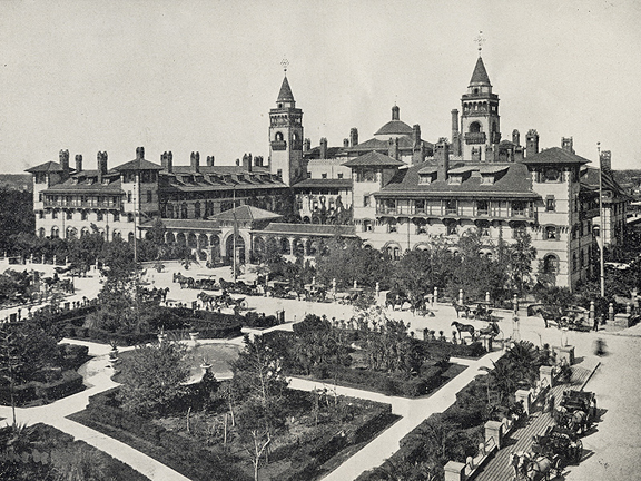 A black and white photograph of the Hotel Ponce de Leon on King Street in St. Augustine, Florida. It is a massive building with many architectural features and windows. The sandy streets in front of it are filled with horse-drawn carriages of different sizes. Also visible is the courtyard of the Alcazar Hotel (now the Lightner Museum), which has a fountain in the center and is filled with shrubbery.