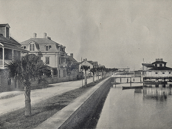 A historical photograph in black and white of the sea wall on Matanzas Bay in St. Augustine, Florida. A sand road parallels the sea wall on the left and is populated by large houses. In the background over the bay, the old St. Augustine Bathhouse stands,
