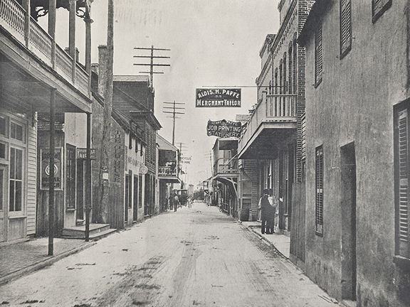 A black and white photograph of a street in St. Augustine, Florida, late 1800s, early 1900s. The street is sand / dirt and spotted with horse droppings. Store signs hang above the street and power lines can be seen above the rooves of the lane of buildings, which are brick, stucco, and wood. Some people can be seen in the background, running errands.