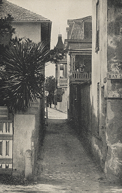 A historical photograph in black and white of Treasury Street in St. Augustine, Florida. Being considered the narrowest street in America, Treasury street appears tight with coquina houses on both sides. Through the alleyway, the Matanzas Bayfront and palm trees are visible.