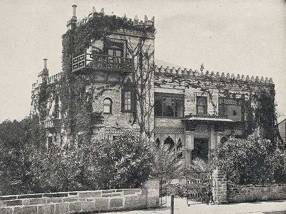 A historical photograph in black and white of the Villa Zorayda on King Street in St. Augustine, Florida. It is a coquina structure meant to mimic the Real Alhambra in Spain. It is a vine covered building with many differently shaped windows and a front garden full of shrubbery.